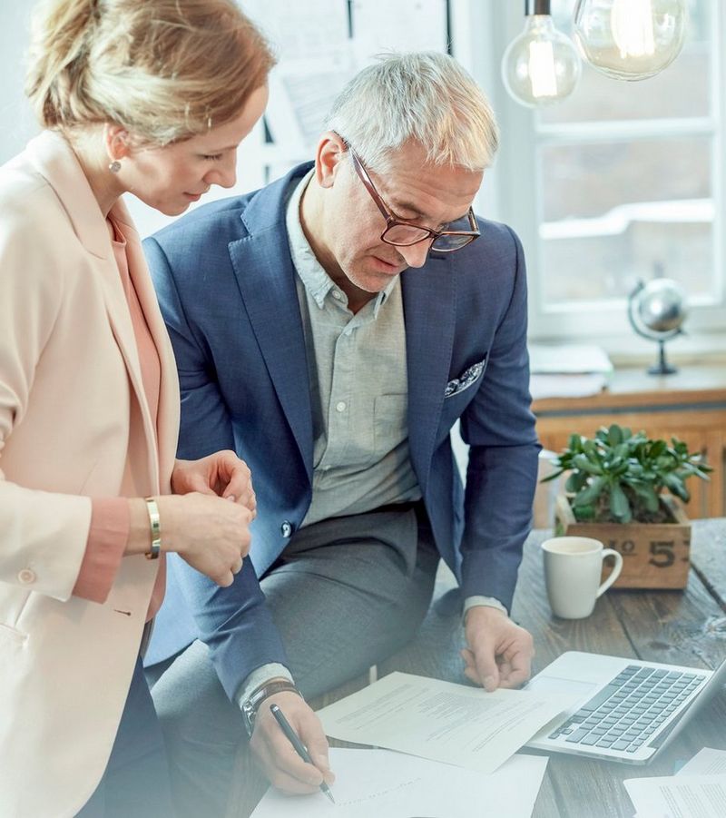 Suited man perched on a rustic table beside a woman wearing a pink jacket writes on a sheet of paper next to an open laptop.