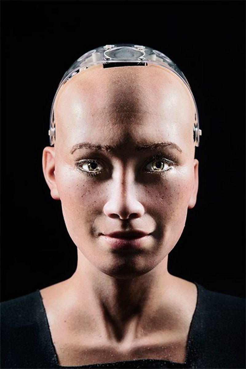 A close-up portrait of Sophia's face and shoulders, with her smiling slightly and her robot skull exposed slightly on top, shot against a black background.