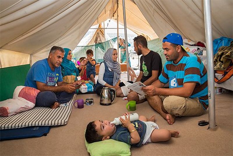 Around 8 relatives sit inside a tent, playing, texting and talking.