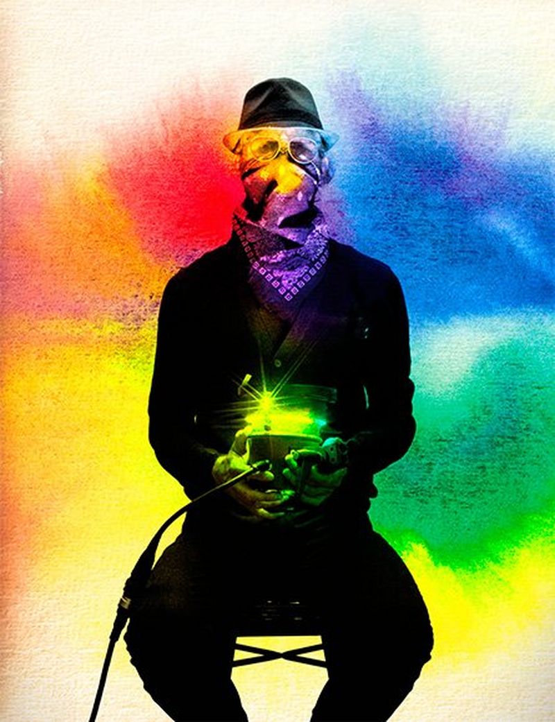 Against a background a splashes of bright colour, a man, dressed in black, sits on a chair. He wears sunglasses and a handkerchief across his nose and mouth, and holds a light source.