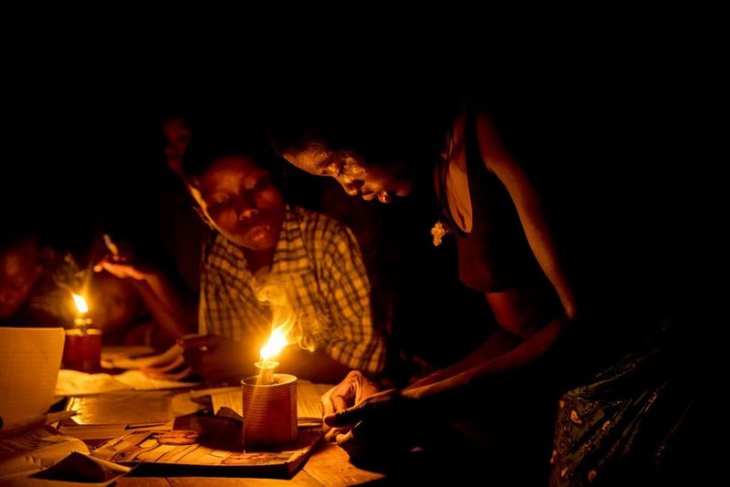 A woman and boys stand over a table of books and study materials, lit by oil lamps.