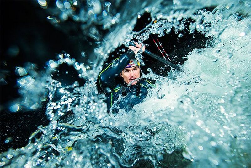 Kayaker Peter Kauzer is seen through a splash of water, as he leans to one side and paddles through the white water, at night.