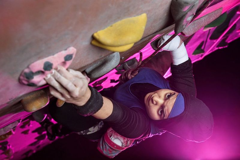 British para climber Anoushé Husain climbs on an indoor climbing wall, lit by a pink light from below.