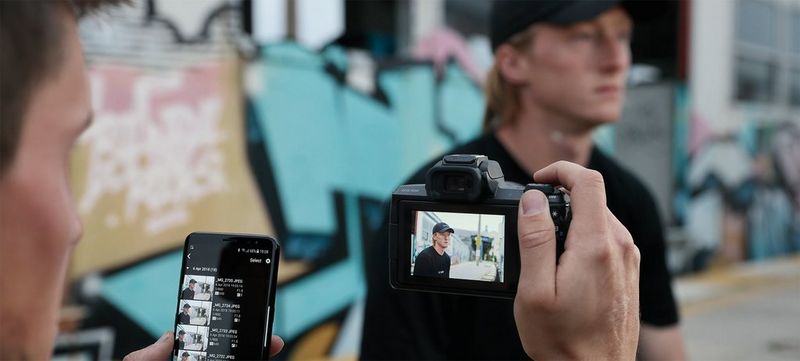 A man in a baseball cap sits in front of a wall with graffiti on. Another man takes his photo.