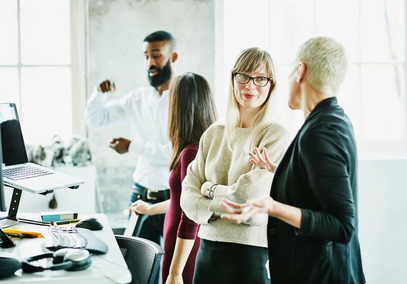 Four office workers stand in pairs chatting, with two gesturing with their hands.