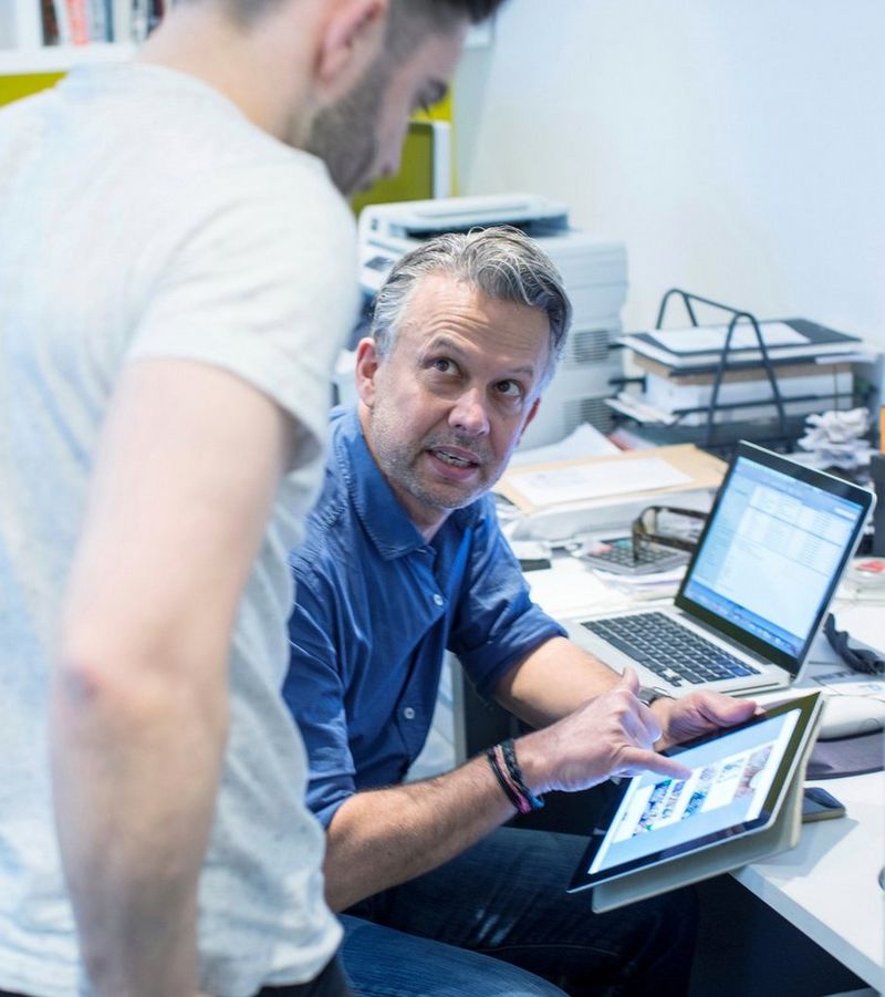 Man working at a laptop turns to another man looking over his shoulder to point out something on a tablet.