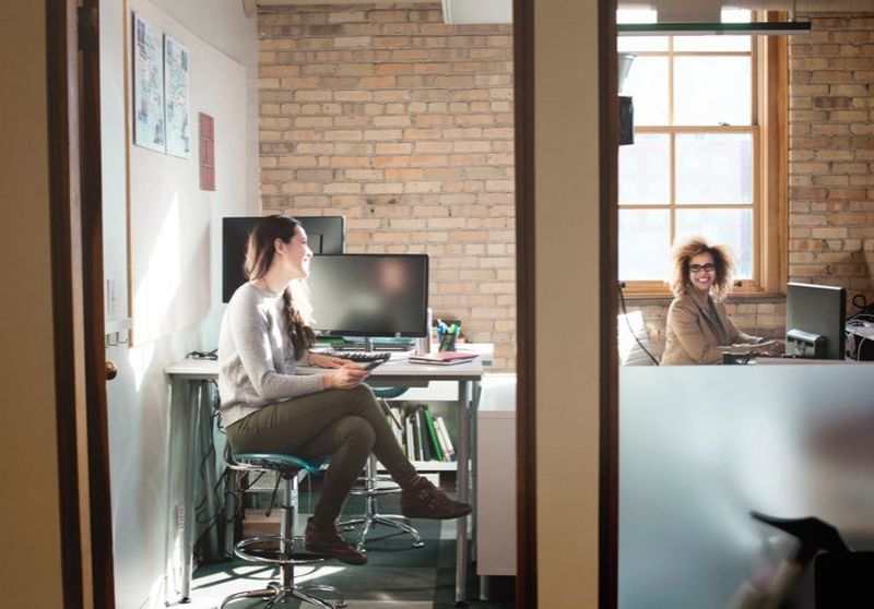 Woman holding a mobile phone and sitting cross-legged at a desk next to a PC chats to a colleague across the office.