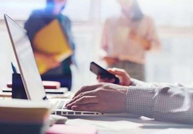 Close-up of a man resting his left hand on a laptop keyboard while holding a mobile phone in his right hand, with out of focus background.