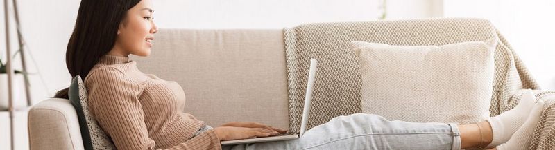 A woman in jeans and a sweater, lies on a beige couch, using a laptop.