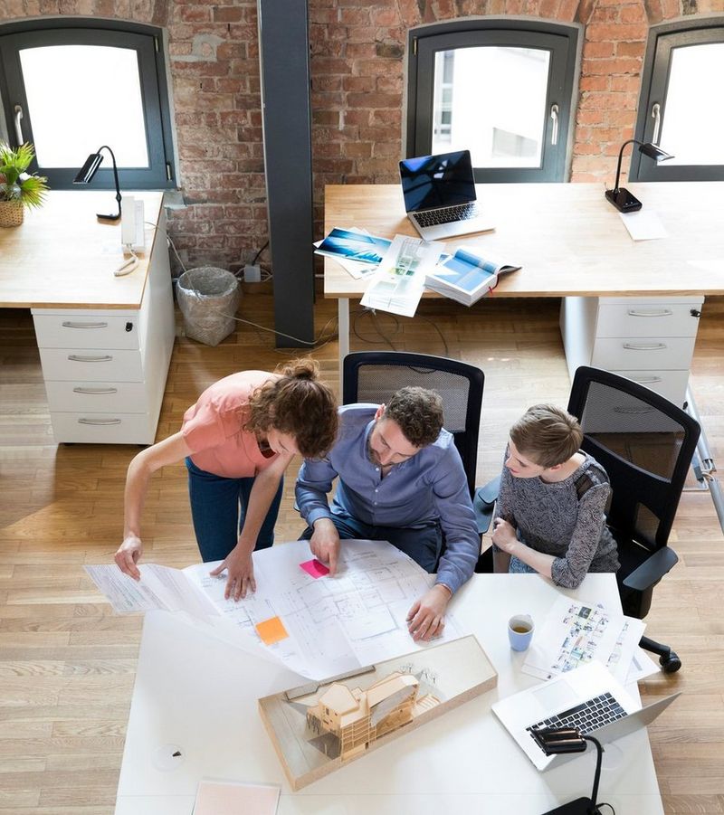 Overhead shot of two women and a man examining architectural drawings, with a model of a building, coffee mug and open laptop on the table.