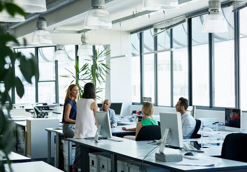 Two women perch on long white desks alongside computers as they chat to three colleagues sitting in front of them.