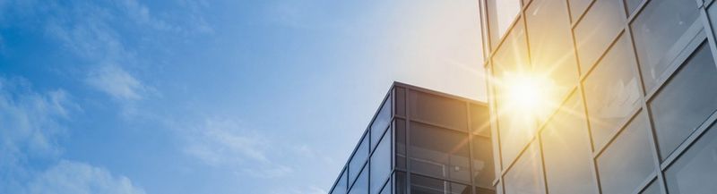 A glass office building on the left, against a blue sky with light clouds. The sun reflects brightly off the building.