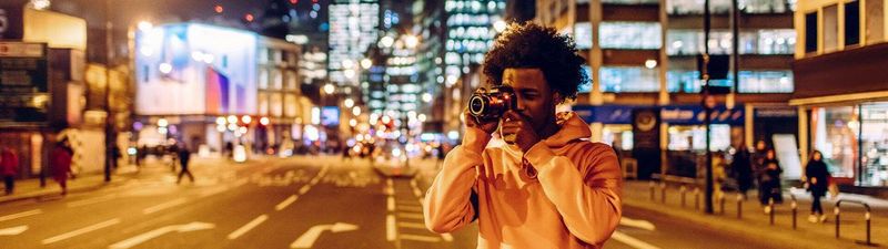 A man stands under a city centre bridge beside a road to take a photo with a Canon camera.