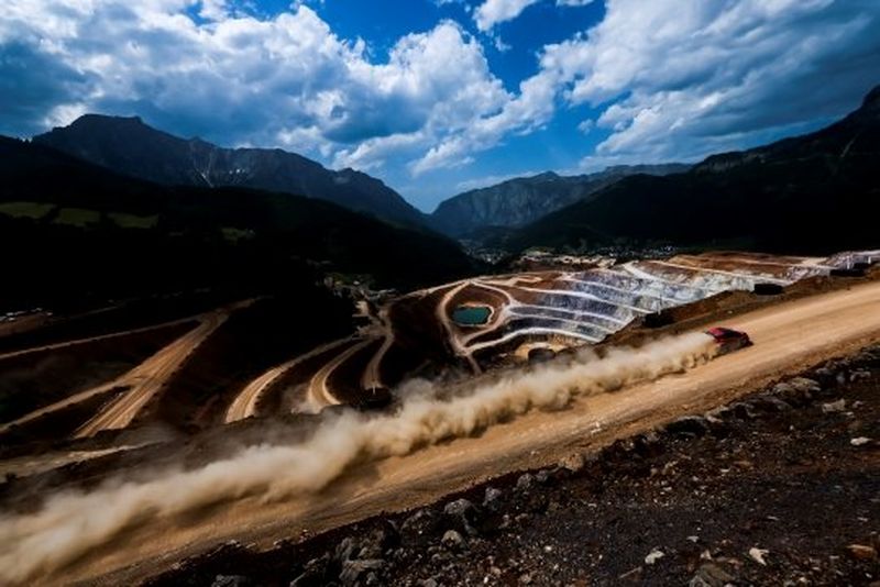  A red rally car kicks up a long trail of dust as it travels at speed along a dirt road above a quarry.