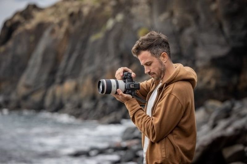 A man in a hooded brown top filming with a D218 camera on a rocky shoreline.