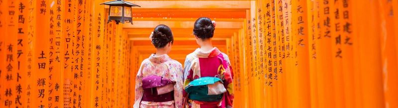 Two geisha stand in front of a red wooden tori gate at fushimi inari shrine in Kyoto, Japan