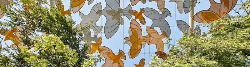 Cut out orange and grey shapes of birds, against a blue sky, with trees on either side. This is the view from beneath of a section of the Terra Sustainability Pavilion at Expo Dubai 2020.