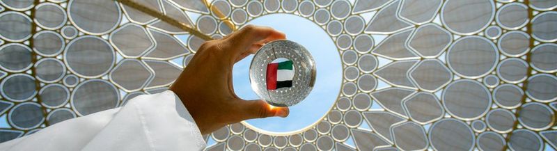 A white sleeved arm holds a glass ball against the sky light through a circular hole in a white ceiling of geometric design. In the centre of the glass ball, the flag of the UAE can be seen.