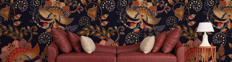 A brown leather sofa and cushions with an occasional table and a lamp to the right. Behind is wallpaper in a repeating design of orange, black and brown leaves and flowers in a modern brocade style, often seen in furnishings and wall coverings.