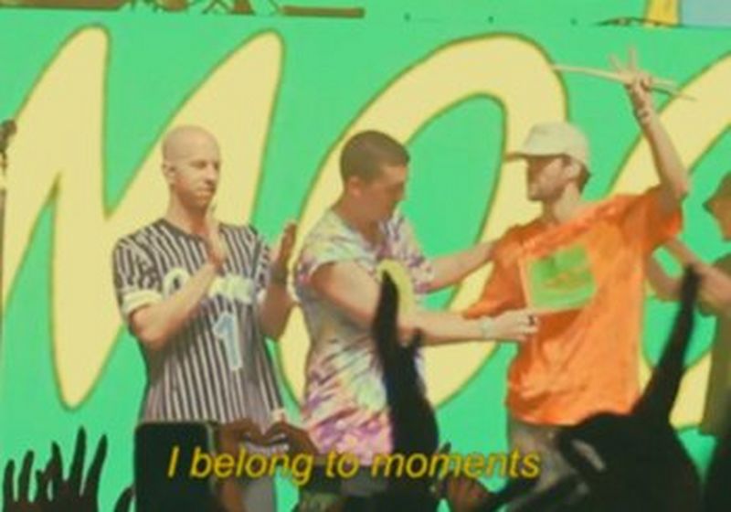 Three men on stage at a festival, with the audience's hands raised in silhouettes in front of them. A caption reading 'I belong to moments' sits at the bottom of the image.