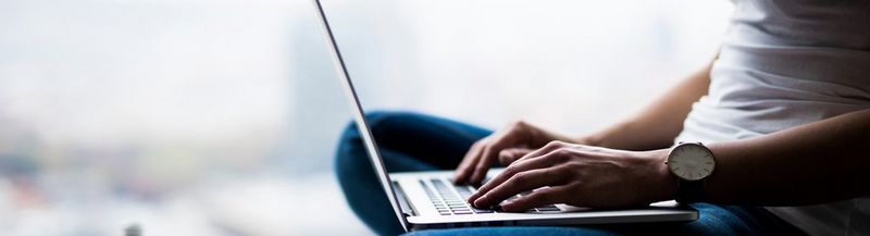 A person in a white t-shirt and blue jeans sits cross-legged, typing on a laptop. Only legs, arms and torso are visible.
