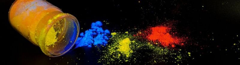 On the left is a small glass bottle on its side. It contains bright yellow powder. Beside it to the right are small piles of bright blue, yellow and red powder. This is all against a black background.