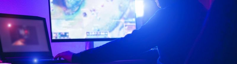 A blue/purple image of a gamer, photographed from the side and behind. They sit in a high backed chair, facing two computer screens.