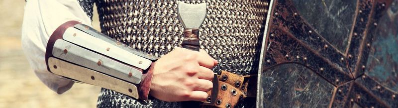 A knight’s chain-mailed torso and arm, with the handle of a sword and partial shield showing.