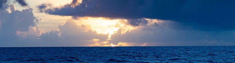 The sun shines from behind clouds above a dark blue ocean (© Richard Garriott de Cayeux, using Canon EOS R / Canon RF24-105mm F4 IS USM)