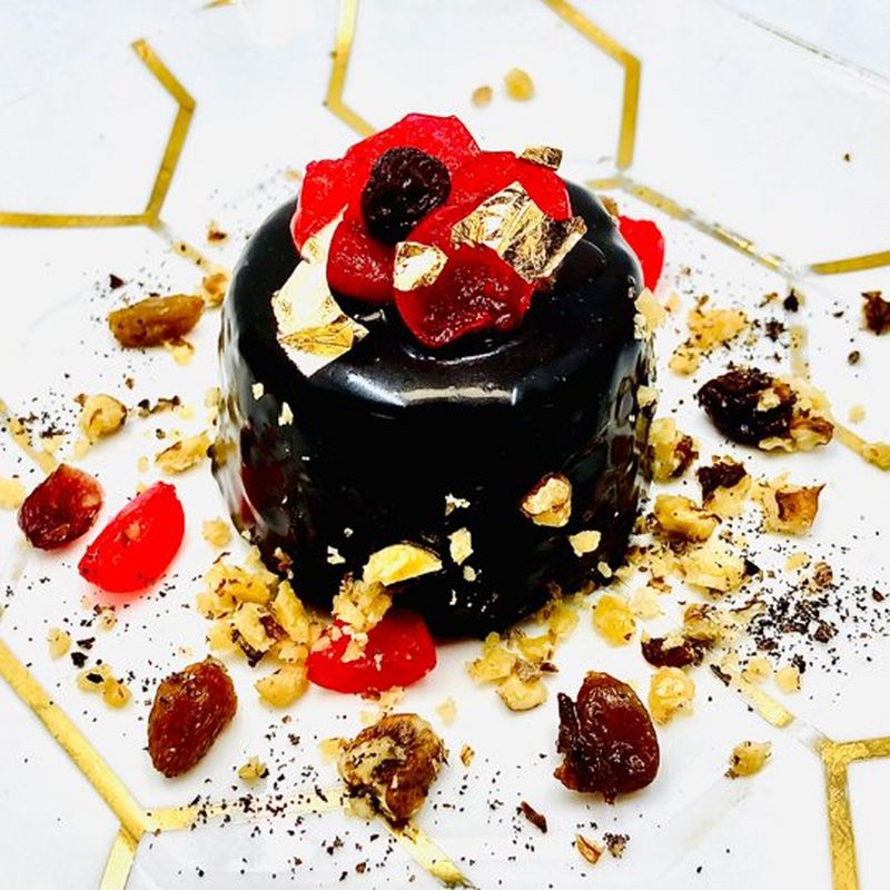 A dark brown chocolate dessert that shines under the light is topped with gold flakes, berries and nuts. It sits on a white plate with a geometric gold honeycomb design.