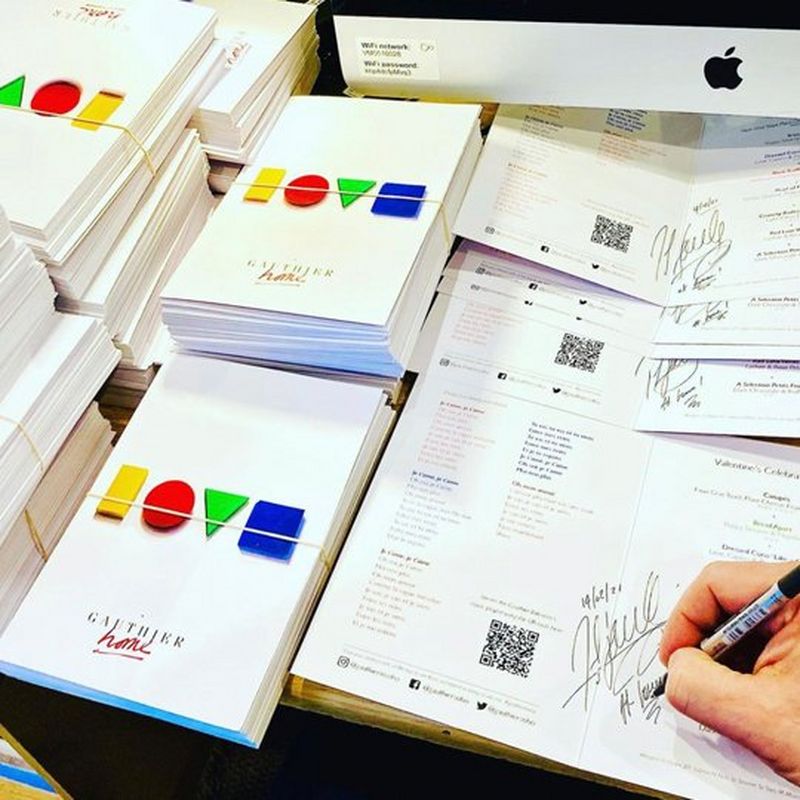 A stack of menus with ‘love’ designed in colourful letters on the front. On the bottom right is a hand holding a pen, signing a menu.