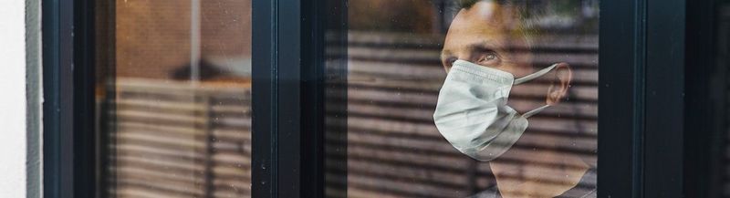 A man wearing a non-surgical face covering and a grey t-shirt, looks out of the window, towards the sky. The glass reflects the outside world, other houses, trees, a fence and the sky.
