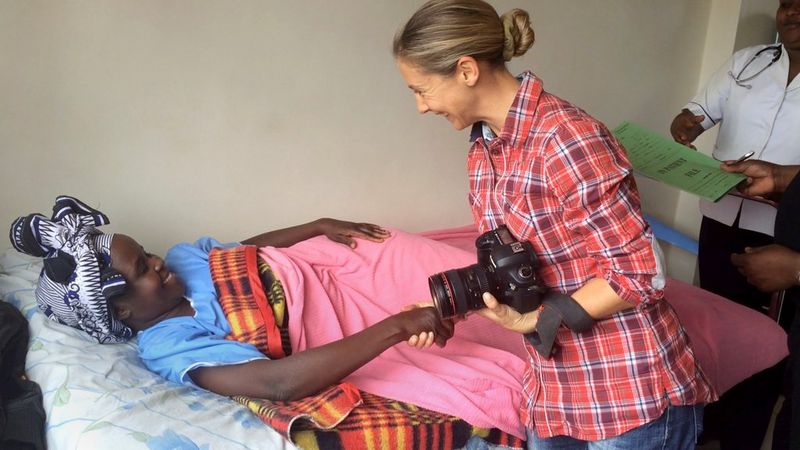 Photographer Georgina Goodwin, holding her Canon camera, greets a woman in a hospital bed in Kenya.
