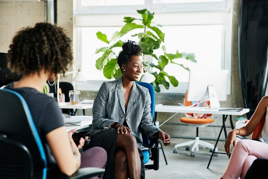 Woman sits on blue chair in front of a plant and talks to two people