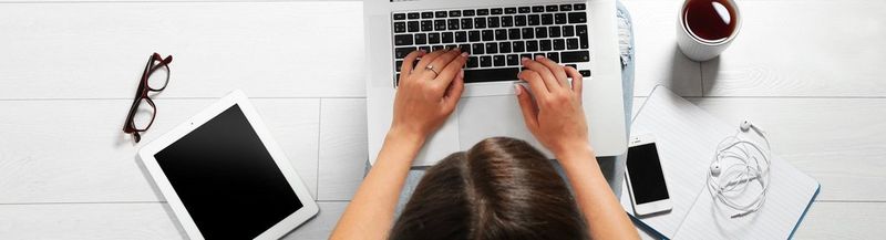 A person viewed from above sits on a white tiled floor with a laptop on their lap. Beside them is a pair of glasses, a tablet, a phone, a notepad, some headphones and a cup of coffee. All white.