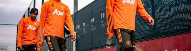 Three footballers walk in line, in front of a black and red barrier. Only one of their faces is shown. They are wearing bright orange shirts with ‘AON’ printed on the front and black shorts with orange stripes down the outer leg. Photo courtesy of Manchester United FC.