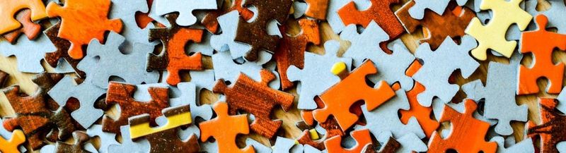 The pieces of a jigsaw puzzle, orange and brown pieces, mixed up with others that are face down, showing only the grey cardboard on their reverse.