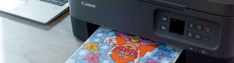 A black Canon printer sits on a table next to a laptop in partial view. On the output tray is a brightly coloured print of an illustration of a woman with orange hair and four eyes, surrounded by colourful birds that have eyes on their wings.