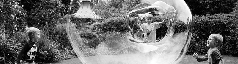 Three children in a garden, one is making a huge soap bubble from a hoop. The others are laughing and screaming with the fun of it all.