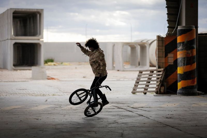 A man in an empty industrial space balances on the back wheel of the BMX he is riding.