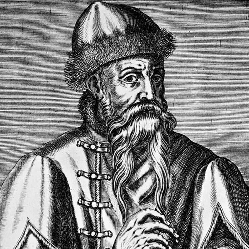 A black and white engraving of Johannes Gutenberg in which he is depicted with a long beard and what appears to be a high, fur-trimmed hat and matching high-necked jacket. He holds what appears to be a lettered stamp in his left hand.