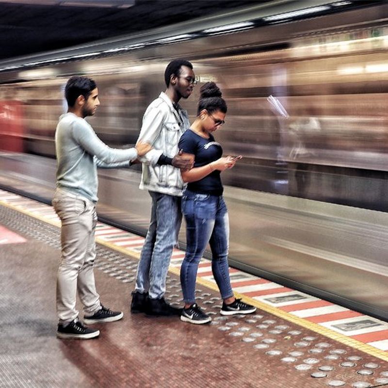 A young woman stands on a train platform as two young men approach her. A train speeds past. 