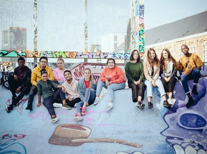 A group of eleven smiling young people, wearing jeans and sweaters, sit at a skate park, posing for the camera.