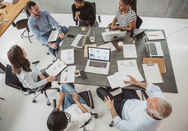 Overhead shot of six casually dressed office workers sat around a communal table comparing graphs on laptops and clipboards.
