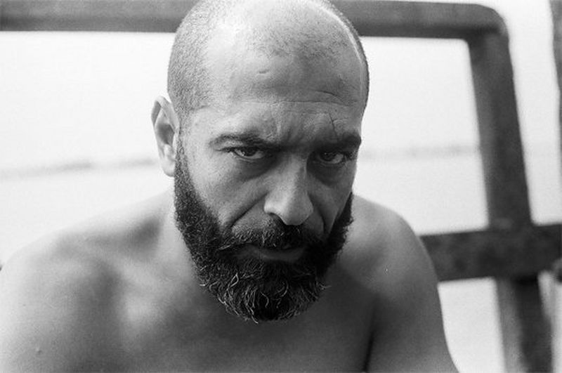 A bald man with a beard looks directly at the camera