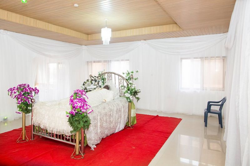 In Ghana, the body of a woman is laid out respectfully on a white bed in a white-draped room with a red rug.