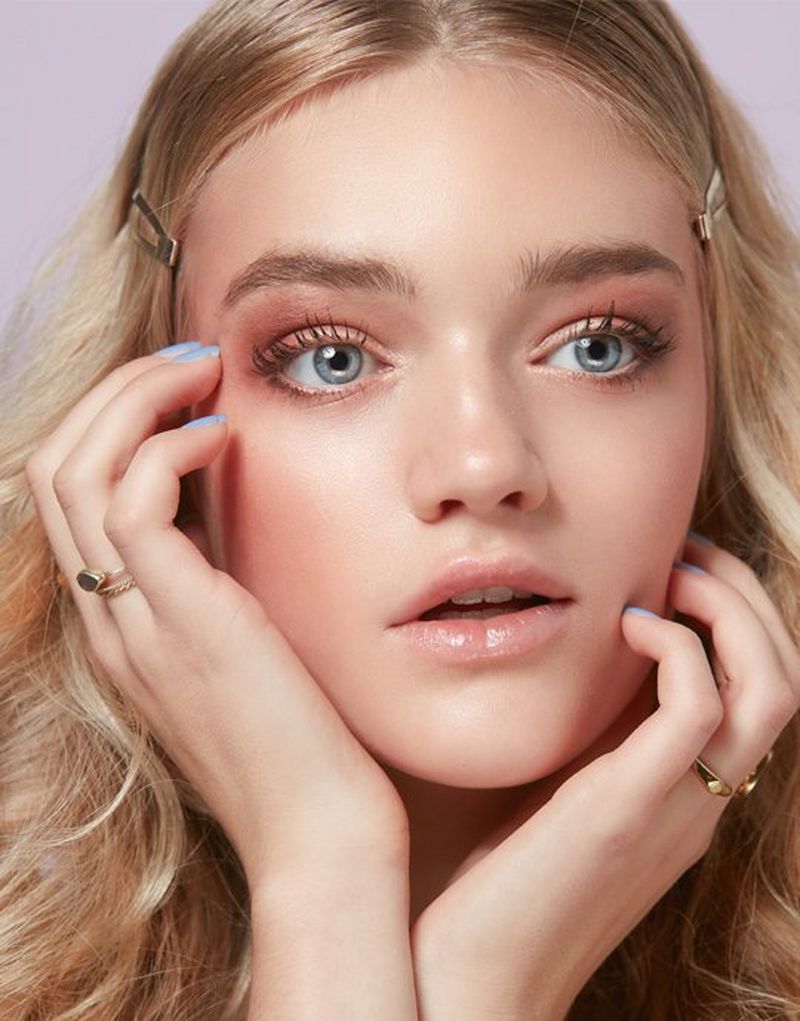 A blonde haired, blue-eyed model wears hairclips to part her hair away from her face. She holds her face in her hands, which have pastel blue nails. Her eye make-up is in gentle shades of rose and she wears a natural lip gloss.