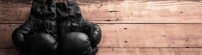 A pair of old-fashioned black lace-up boxing gloves, lying against a wooden background.