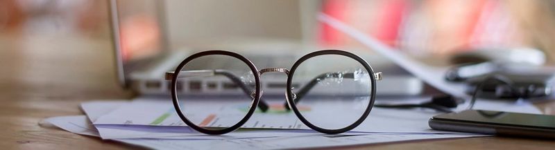 A pair of spectacles sat on top of paperwork, in front of a laptop and next to a phone