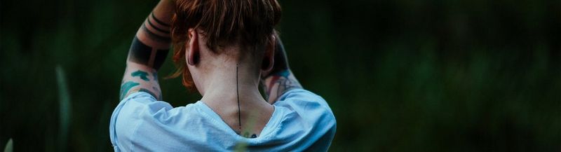 A person in a light-coloured t-shirt, photographed from the back, against a natural green background. They have their tattooed arms held in the air, above their head, as though holding their red hair up. © Marta Sobilo